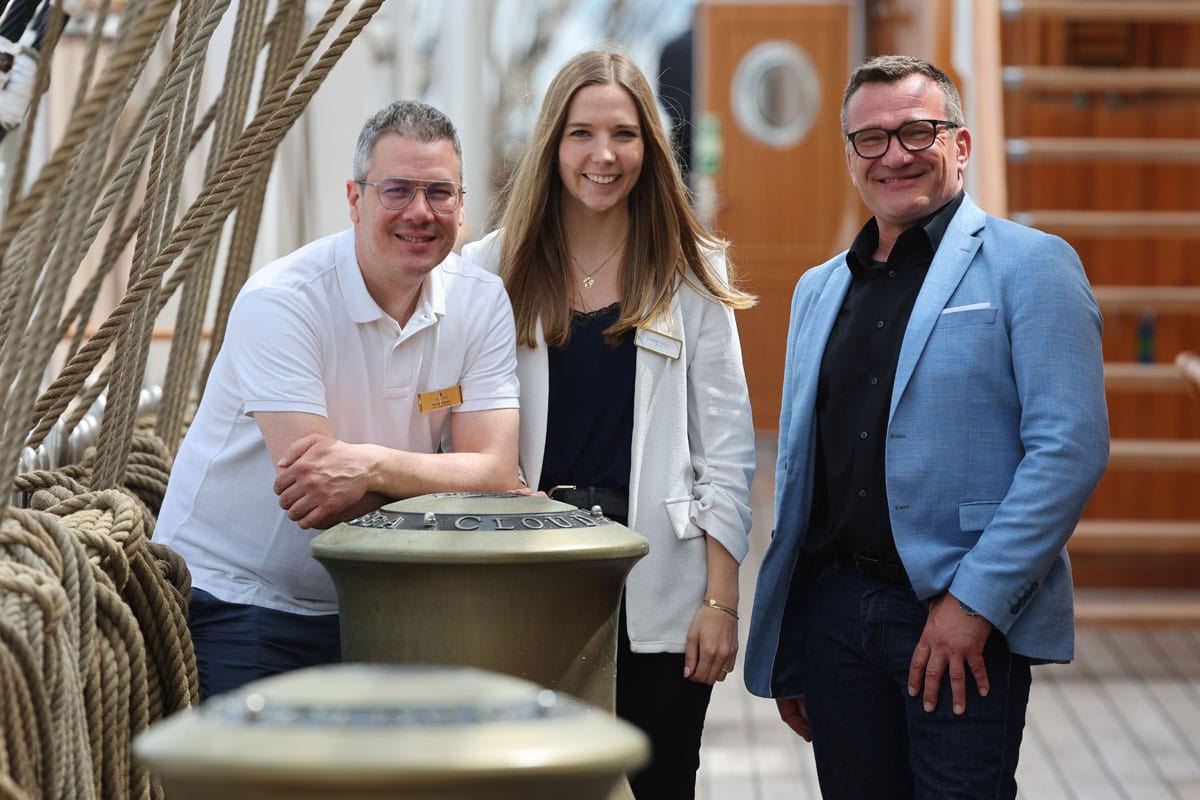 Zwei Männer und eine Frau in legerem Business-Look posieren an Deck eines luxuriösen Großseglers.