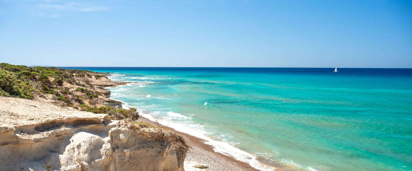 Blick über einen hellen, von Klippen begrenzten Sandstrand aufs Meer, das zwischen Türkis und Dunkelblau changiert. Ein kleines weißes Segelboot ist am Horizont zu sehen. Der Himmel ist blau, die Sonne scheint.