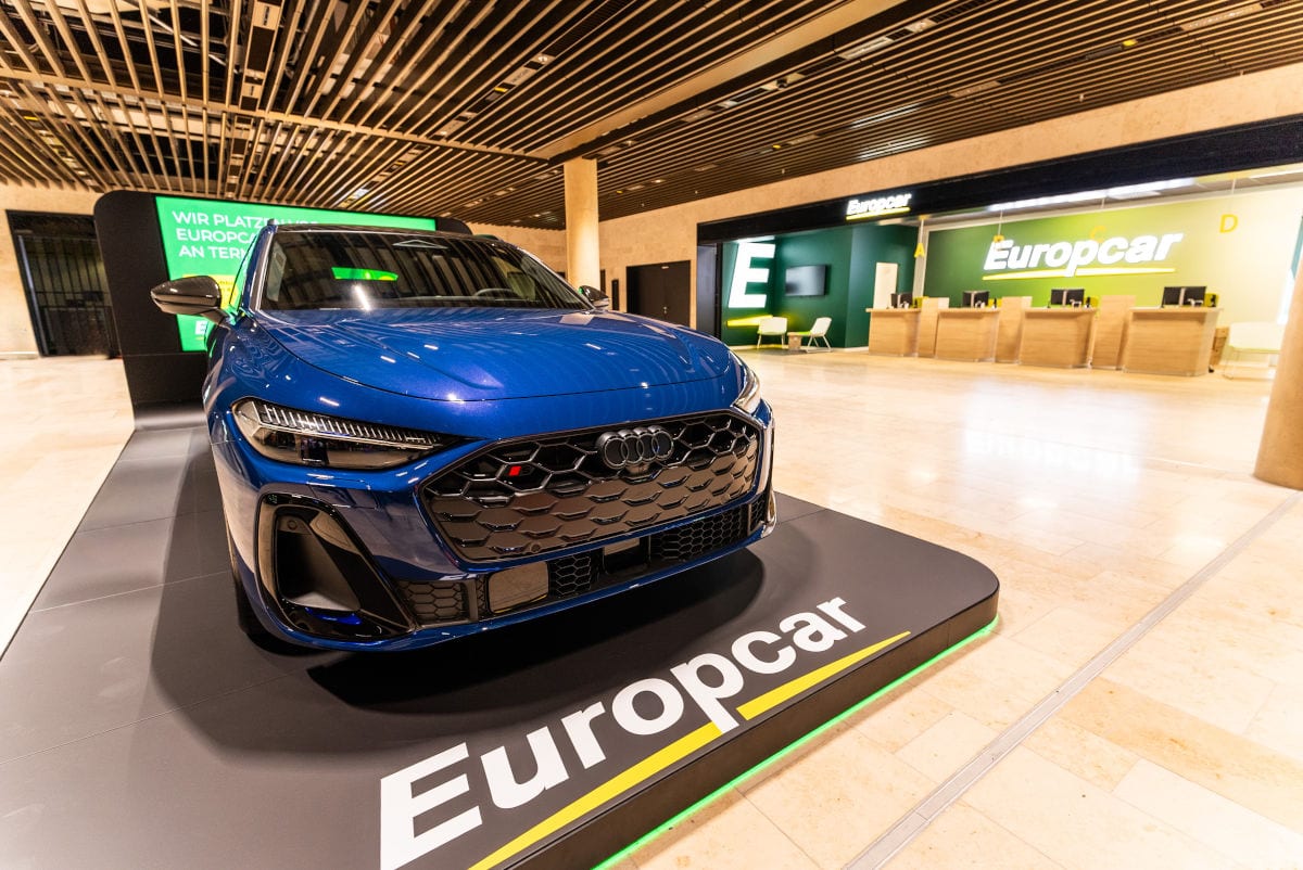 Im Bild ist ein neuer blauer Audi vor dem Europcar-Schalter am Frankfurter Flughafen Terminal 3 zu sehen. Das Auto steht prominent auf einer Plattform mit dem Europcar-Logo, während im Hintergrund die moderne Autovermietung zu erkennen ist. Die Szene vermittelt den Eindruck eines hochwertigen Mietwagenservices am Flughafen Frankfurt.