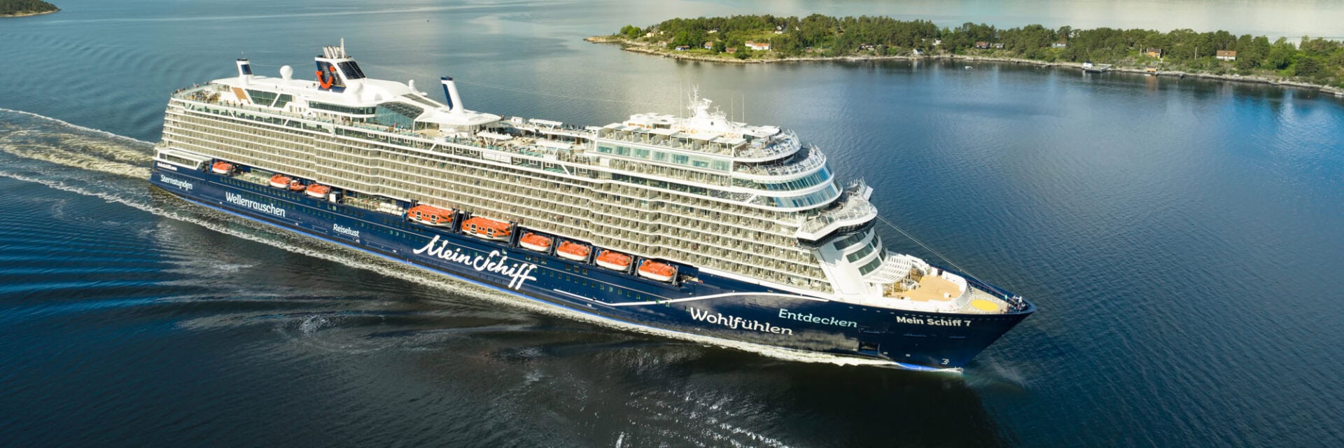 Das Bild zeigt das Kreuzfahrtschiff Mein Schiff 7 von TUI Cruises, das durch den Oslofjord in Norwegen fährt. Im Hintergrund sind bewaldete Hügel, eine Insel mit Häusern und der ruhige Fjord unter blauem Himmel mit Wolken zu sehen.