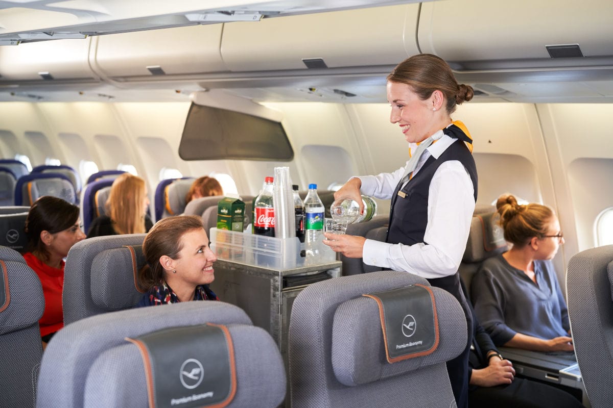 In einer Lufthansa Economy Kabine serviert eine Flugbegleiterin Getränke mit einem Bordservicewagen. Mehrere Passagiere sitzen entspannt in ihren Sitzen, während Wasser und Softdrinks wie Cola angeboten werden.