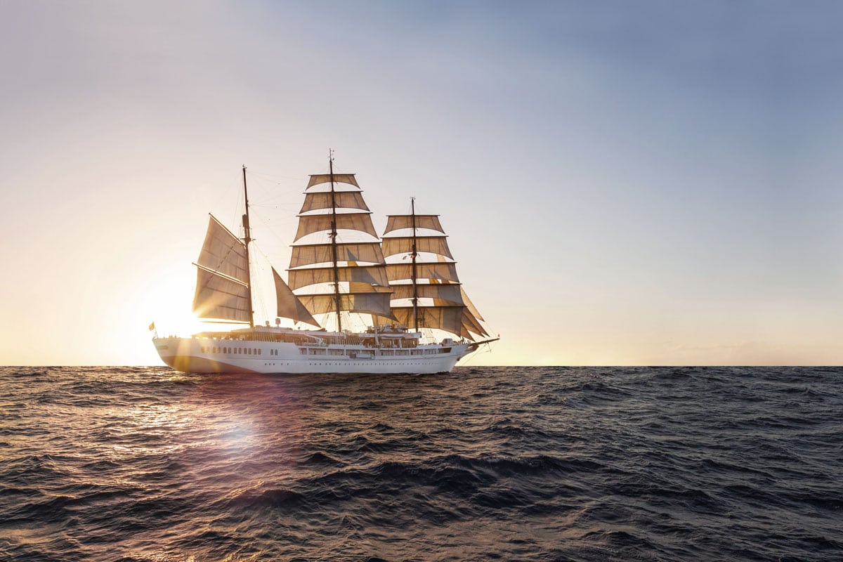 Ein Segelkreuzfahrtschiff mit mehreren Masten und weißen Segeln fährt bei Sonnenuntergang über das offene Meer. Die goldene Abendsonne beleuchtet das Wasser und das elegante Kreuzfahrtschiff.