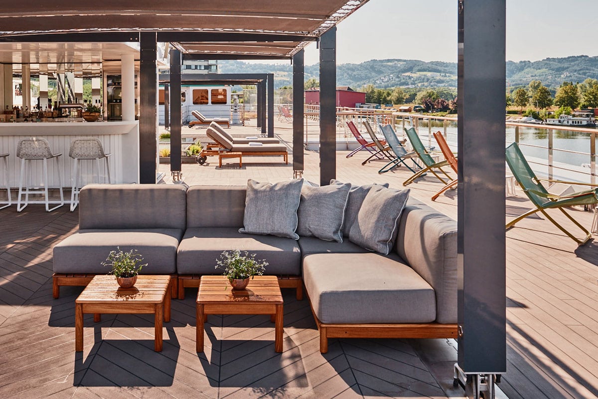 Das Bild zeigt das Sonnendeck eines Flusskreuzfahrtschiffs mit einer stilvollen Lounge-Ecke, einer modernen Bar und zahlreichen Liegestühlen mit Blick auf einen ruhigen Fluss und grüne Hügellandschaft. Die offene Terrasse ist mit gemütlichen Sitzmöbeln und Holzdekor gestaltet und lädt zur Entspannung ein.