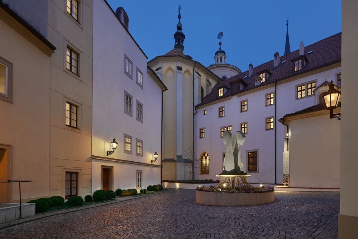 Ruhiger, gepflasterter Innenhof historischer Gebäude bei Abenddämmerung, warm beleuchtet durch Laternen; in der Mitte steht ein moderner Brunnen mit stilisierter Skulptur, umgeben von hellen Fassaden und Fenstern.