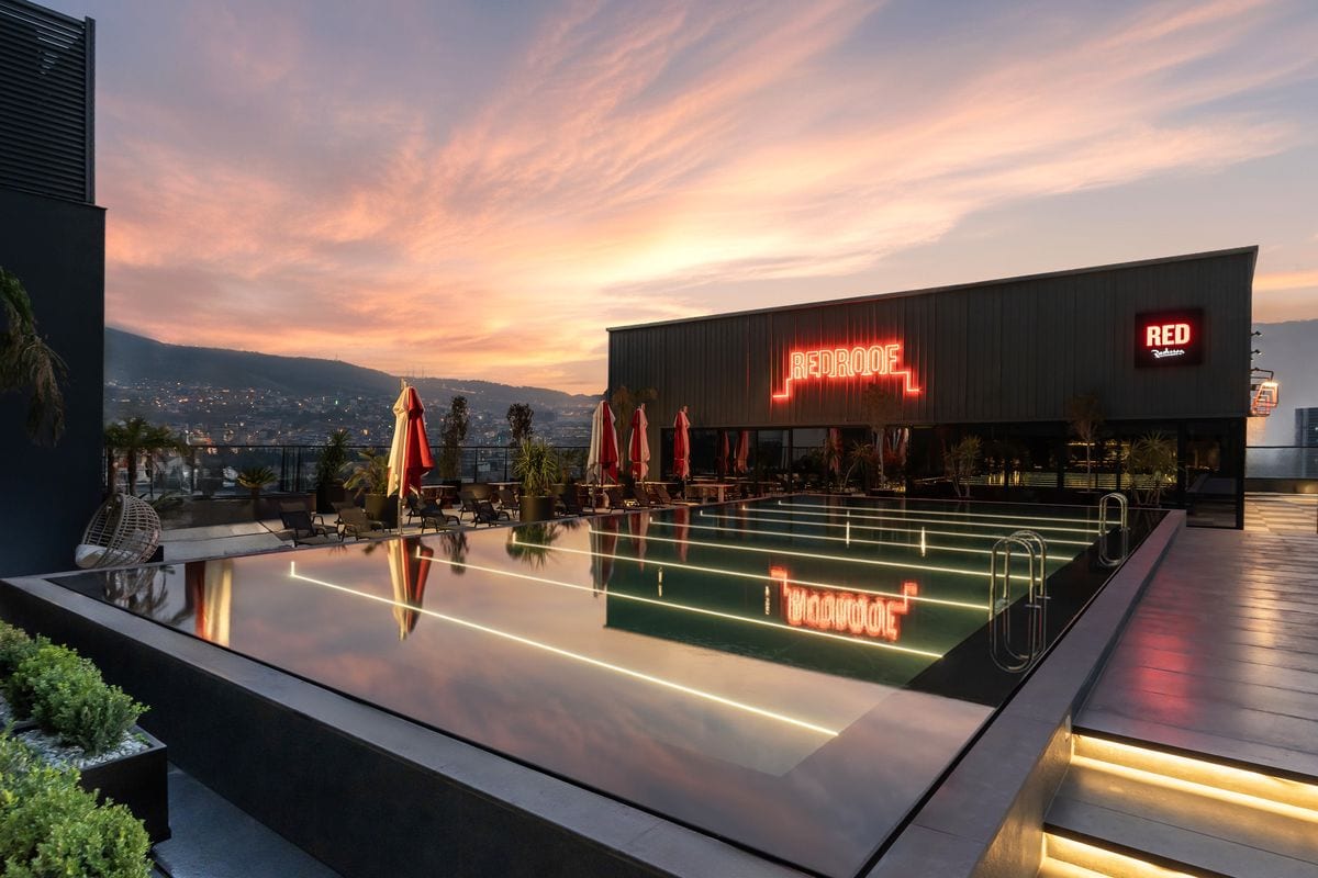 Dachterrasse eines Hotels in Izmir bei Sonnenuntergang mit beleuchtetem Pool, roten Sonnenschirmen, Neon-Schriftzug und Blick über die Stadt.