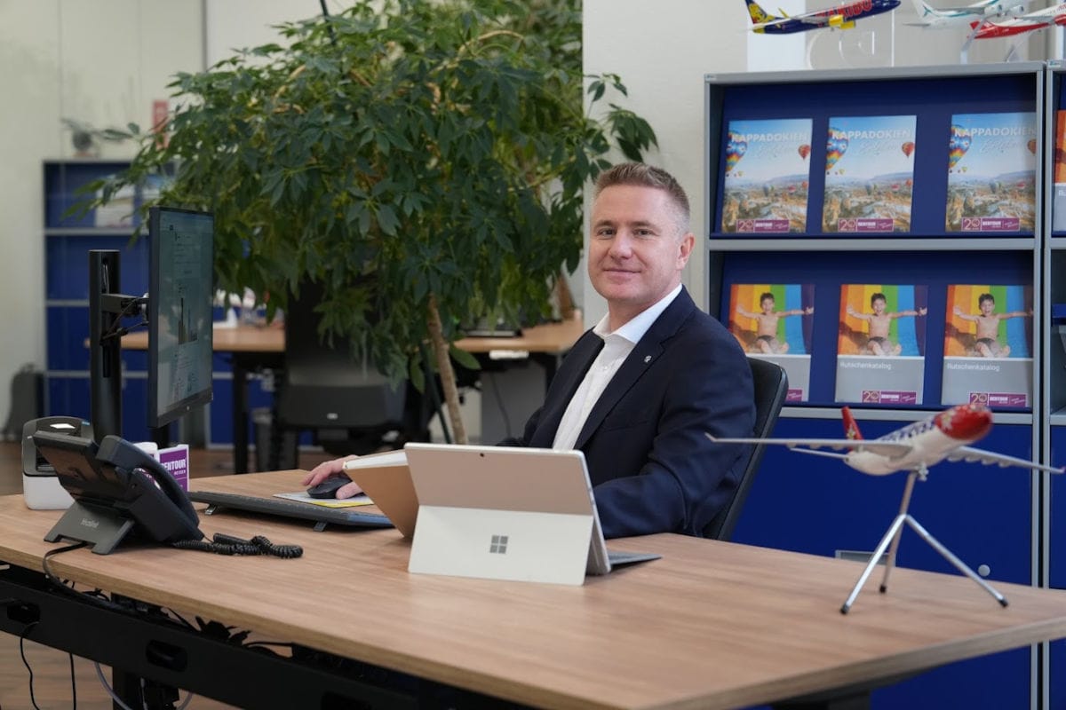 Ein Mann im Anzug sitzt an einem Schreibtisch in einem modernen Büro und blickt in die Kamera. Vor ihm stehen ein Computer, ein Tablet und ein Telefon. Im Hintergrund sind eine große Zimmerpflanze sowie ein Regal mit Reisekatalogen und Modellflugzeugen zu sehen.