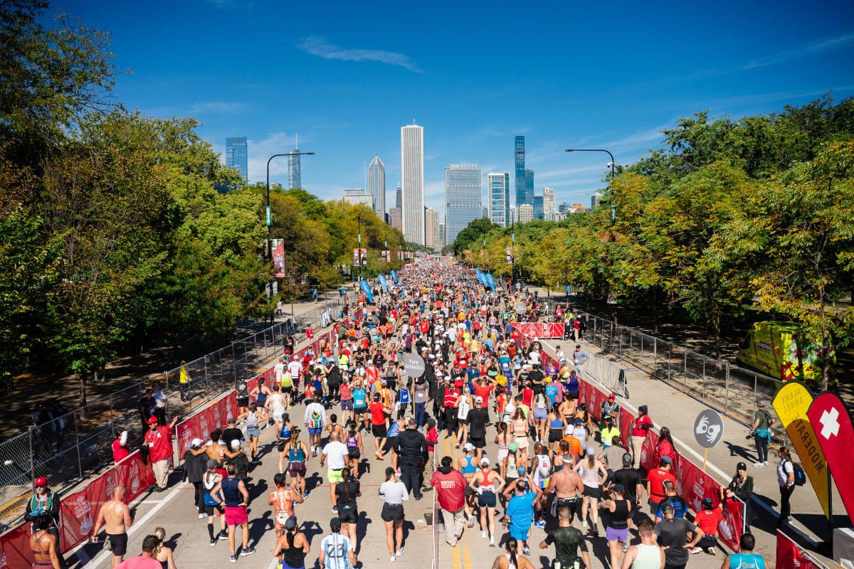 Teilnehmerfeld beim Chicago Marathon: Tausende Läuferinnen und Läufer bewegen sich dicht gedrängt auf einer breiten Straße, gesäumt von Bäumen und Absperrungen, während im Hintergrund die Skyline von Chicago mit Hochhäusern unter blauem Himmel zu sehen ist.