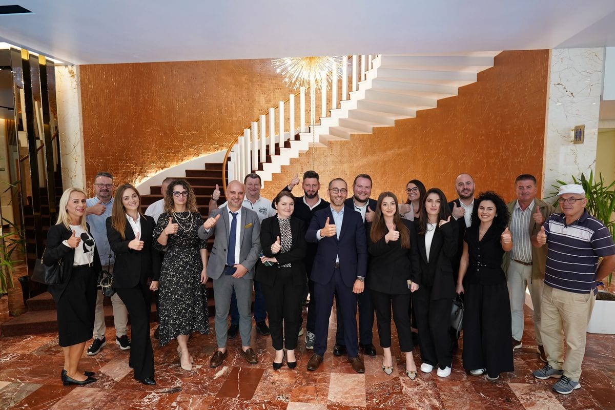 Eine Gruppe von etwa 20 Personen steht in einer Hotellobby vor einer geschwungenen Treppe und blickt lächelnd in die Kamera. Alle zeigen den Daumen nach oben. Die Gruppe besteht aus Männern und Frauen. Die Umgebung wirkt elegant mit marmornem Boden und goldfarbener Wandgestaltung.