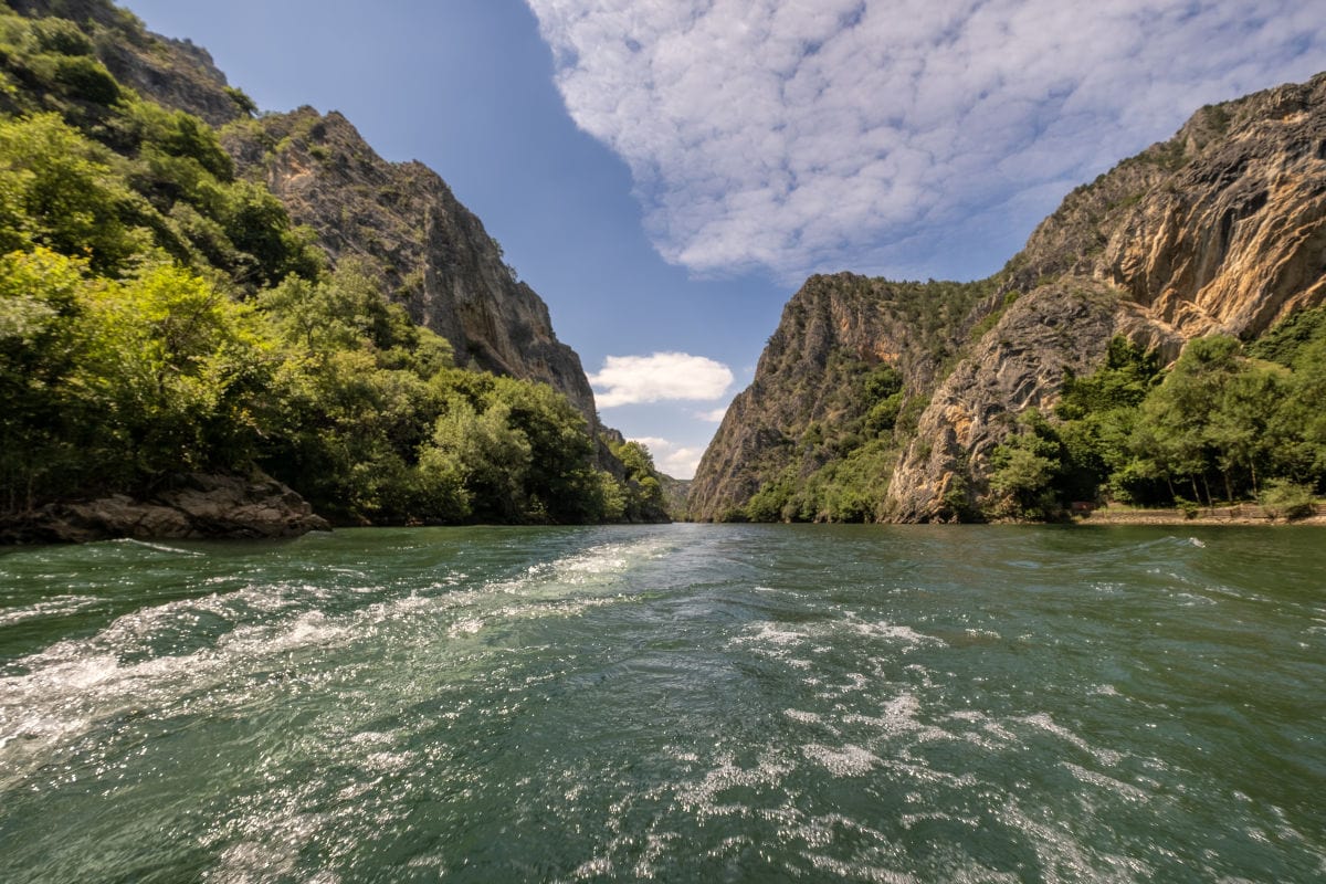 Blick von einem Boot auf einen grünen Fluss, der durch eine enge Schlucht mit steilen, bewaldeten Felswänden auf beiden Seiten verläuft. Das Wasser ist leicht aufgewühlt, und über der Landschaft spannt sich ein blauer Himmel mit lockeren, weißen Wolken.