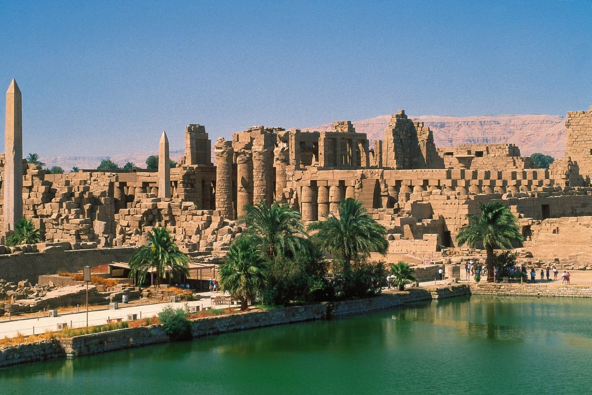Blick auf die Tempelruinen von Karnak in Luxor: sandfarbene antike Säulen und Obelisken, Palmen am Ufer eines Wasserbeckens, dahinter Wüstenhügel unter blauem Himmel.