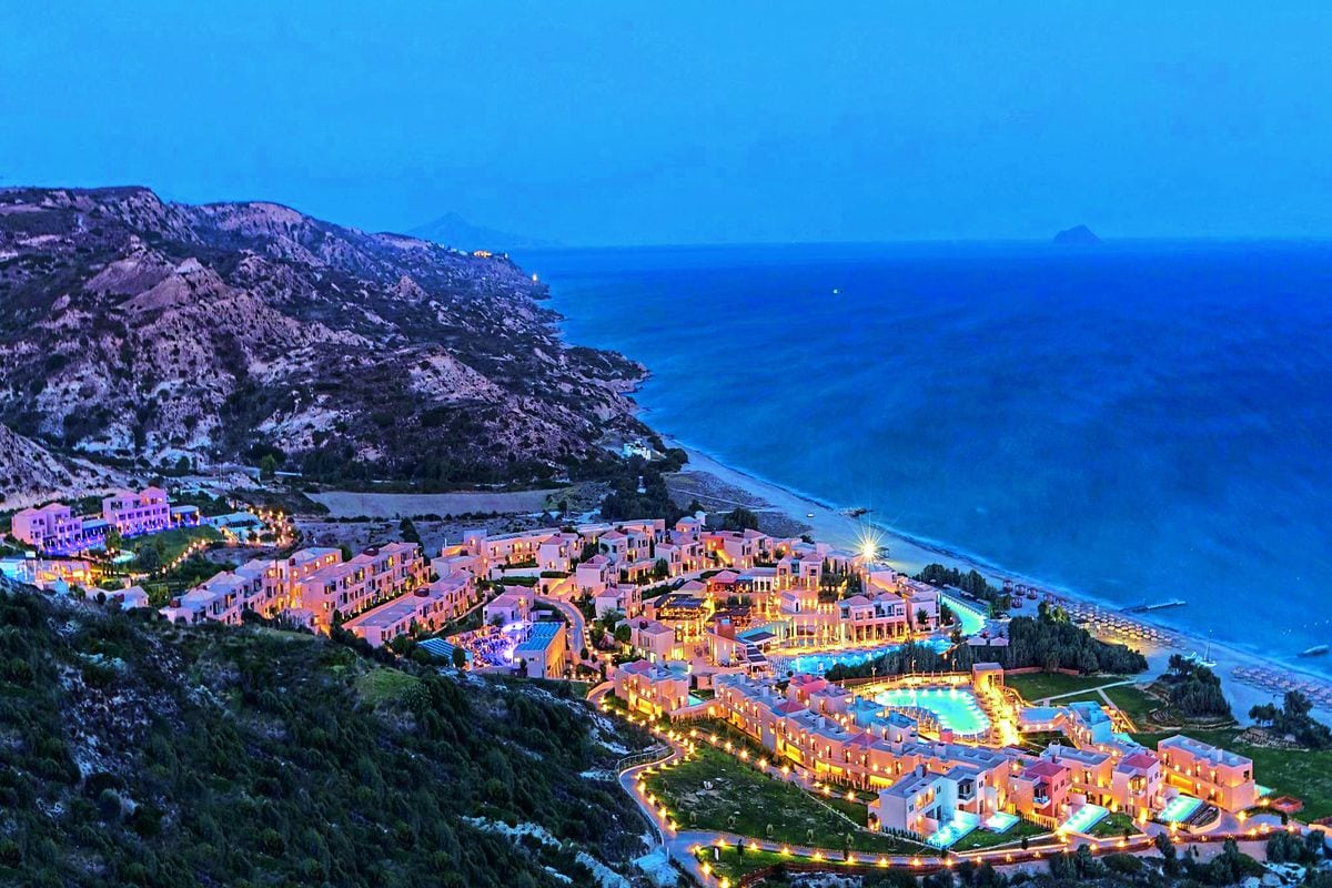 Weitläufige Hotelanlage an einer Küste bei Dämmerung mit beleuchteten Gebäuden, Pools und direktem Zugang zum Meer; im Hintergrund Hügel und das offene Wasser.