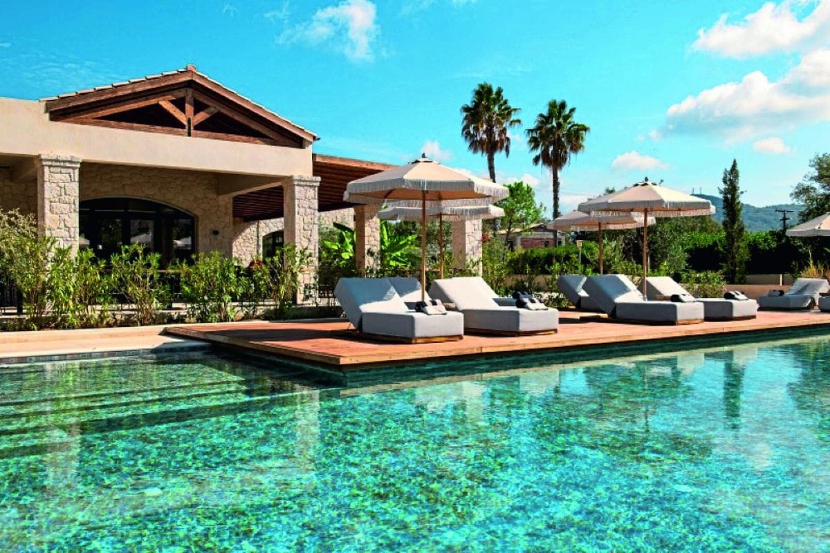 Stilvoller Poolbereich mit flachem, türkisfarbenem Wasser, eleganten Sonnenliegen und Sonnenschirmen vor einem mediterranen Gebäude mit Natursteinfassade und üppiger Gartenlandschaft.