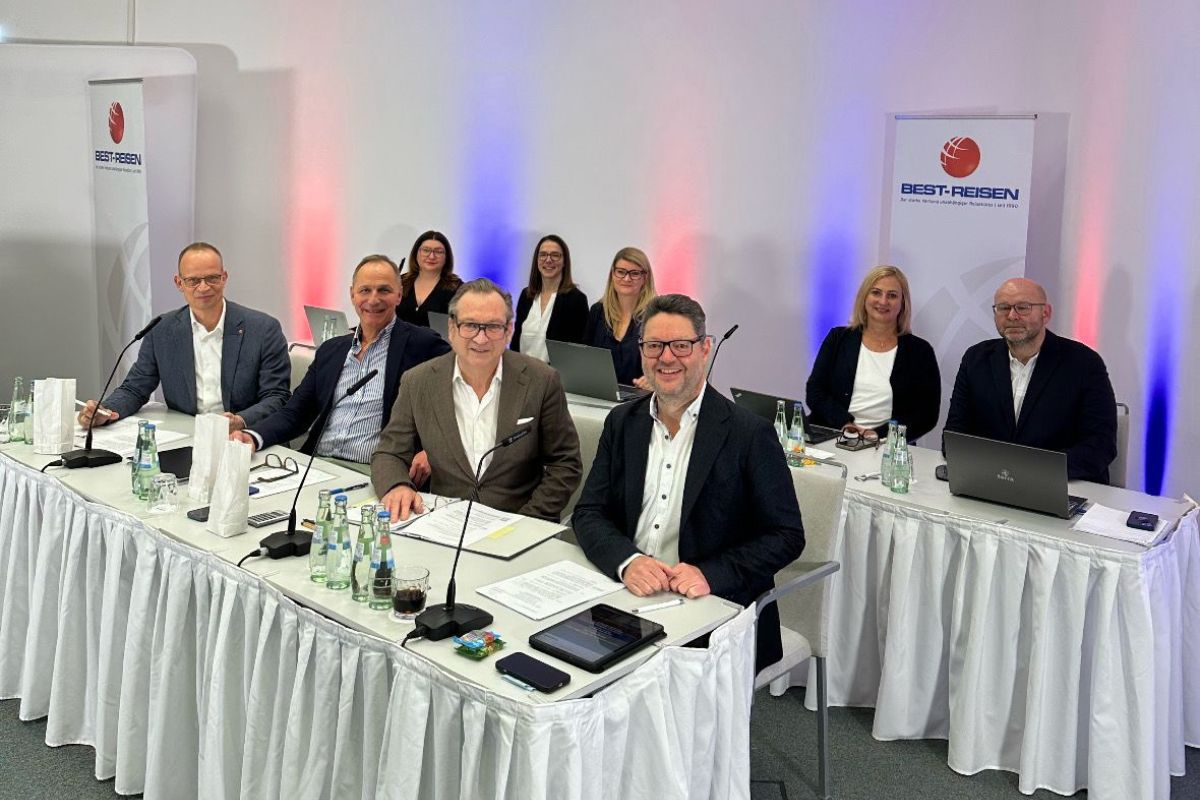 Gruppe von neun Personen bei einer Sitzung von Best-Reisen: Vorne sitzen vier Männer an einem Konferenztisch mit Mikrofonen, Unterlagen und Getränken, dahinter fünf weitere Personen mit Laptops. Im Hintergrund stehen Roll-ups mit dem Best-Reisen-Logo sowie eine Wandbeleuchtung in Rot und Blau.