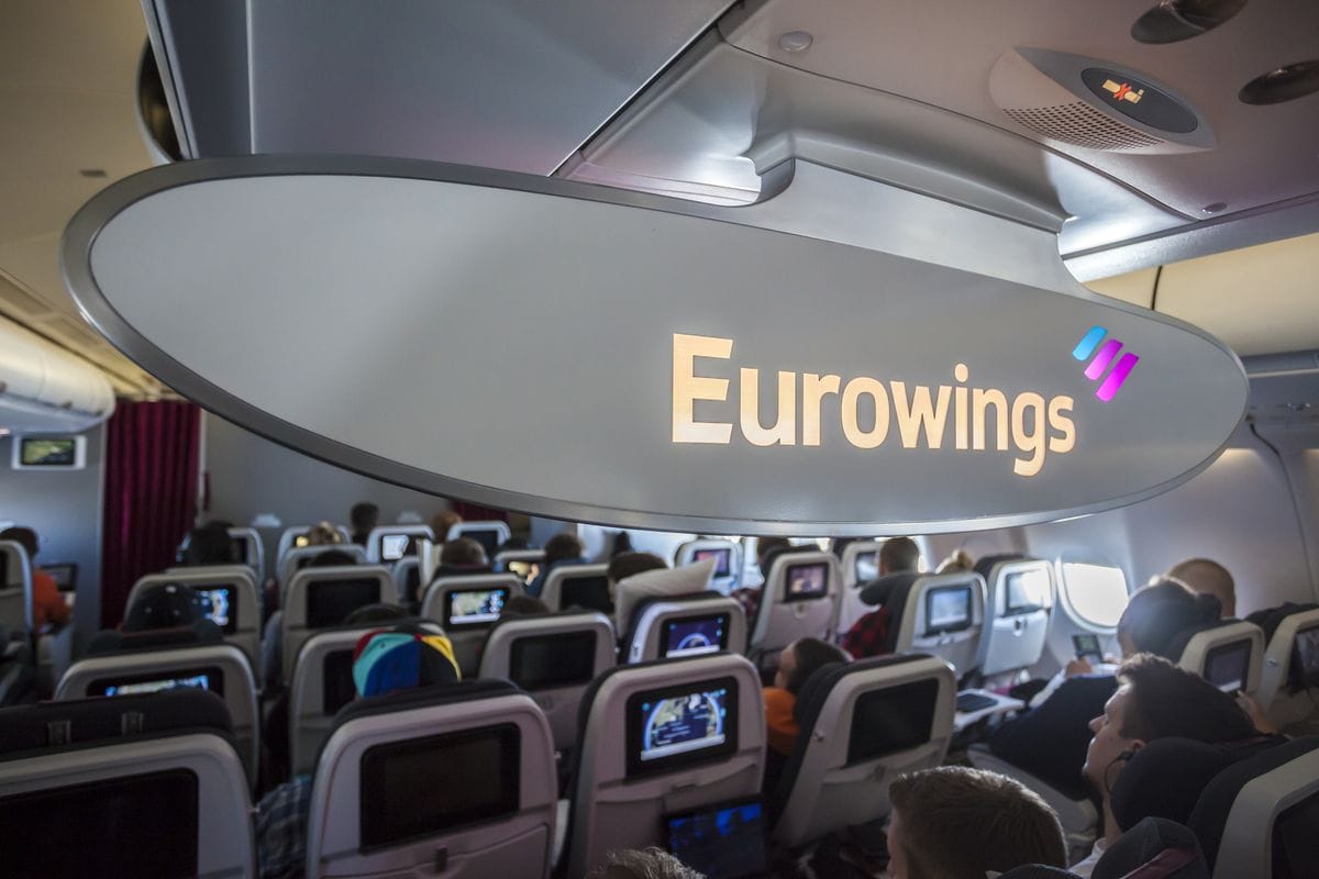 Kabinenansicht in einem Eurowings-Flugzeug mit sitzenden Passagieren und großem Eurowings-Schriftzug im Vordergrund.