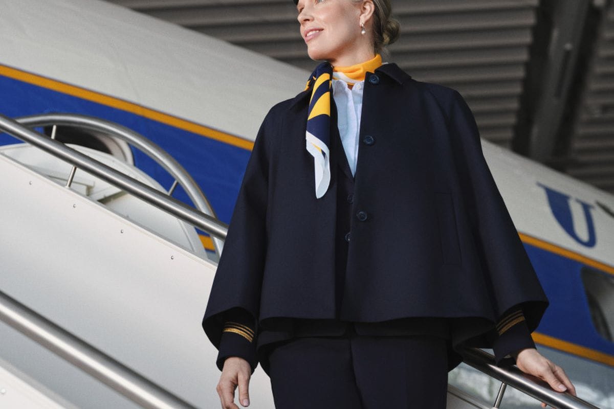 Mitarbeiterin in neuer Lufthansa-Uniform mit dunkelblauem Cape und gelb-blauem Halstuch auf einer Flugzeugtreppe vor einer Maschine.