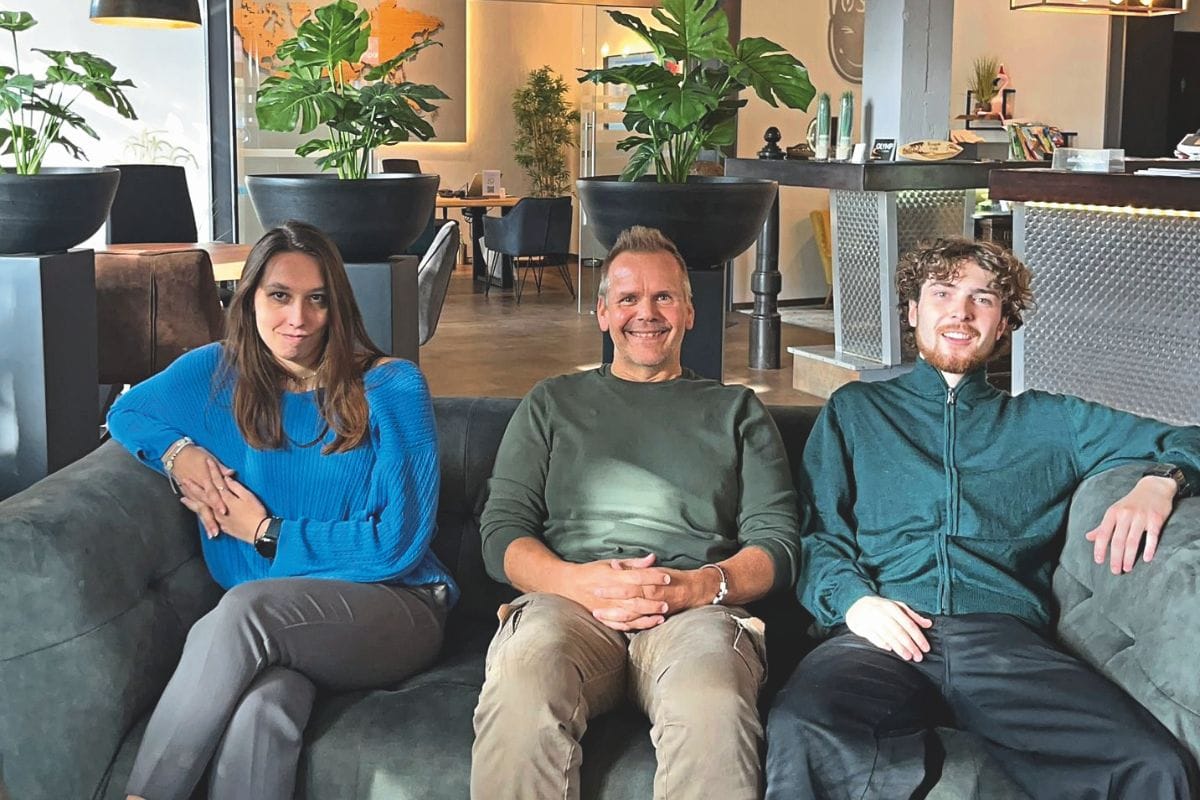 Drei Personen sitzen entspannt auf einem Sofa in einem modern eingerichteten Reisebüro mit Pflanzen und warmem Licht; sie blicken in die Kamera und lächeln, im Hintergrund sind Beratungsbereiche und ein Empfangstresen zu sehen.