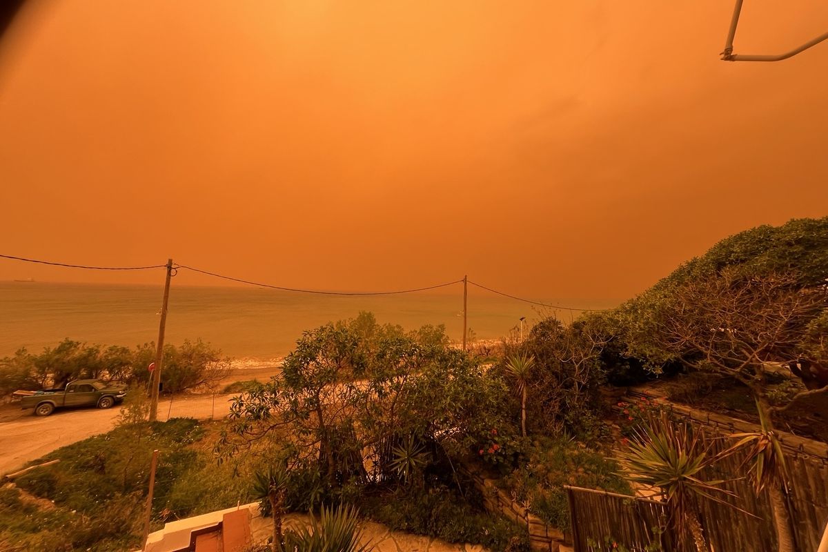 Rötlich verfärbter Himmel über einer Küstenlandschaft auf Kreta: Gebäude, Bäume und Meer sind in dichtem Saharastaub und unheimliches orange-rotes Licht getaucht.