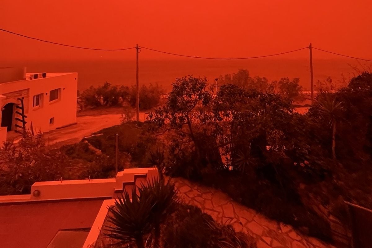 Rötlich verfärbter Himmel über einer Küstenlandschaft auf Kreta: Gebäude, Bäume und Meer sind in dichtem Saharastaub und unheimliches orange-rotes Licht getaucht.
