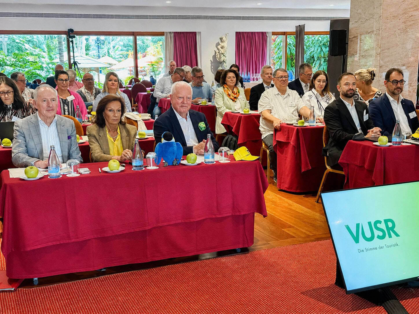 Eine Gruppe von etwa 20–30 erwachsenen Personen sitzt in einem Konferenzraum an Tischen mit roten Tischdecken und hört aufmerksam zu. Auf den Tischen stehen Wasserflaschen, Gläser und grüne Äpfel. Im Vordergrund sitzen mehrere gut gekleidete Teilnehmer in Business-Kleidung. Rechts vorne ist ein Bildschirm mit der Aufschrift „VUSR – Die Stimme der Touristik“. Große Fenster im Hintergrund lassen viel Tageslicht herein und geben den Blick auf eine grüne Außenanlage frei.