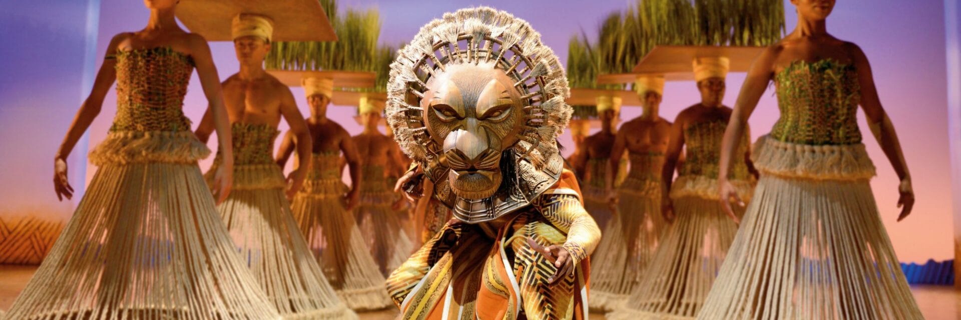 Bühnenbild aus dem Musical „Der König der Löwen“: Darsteller in aufwendigem Löwenkostüm im Vordergrund, begleitet von Ensemble in afrikanisch inspirierten Kostümen und Requisiten.