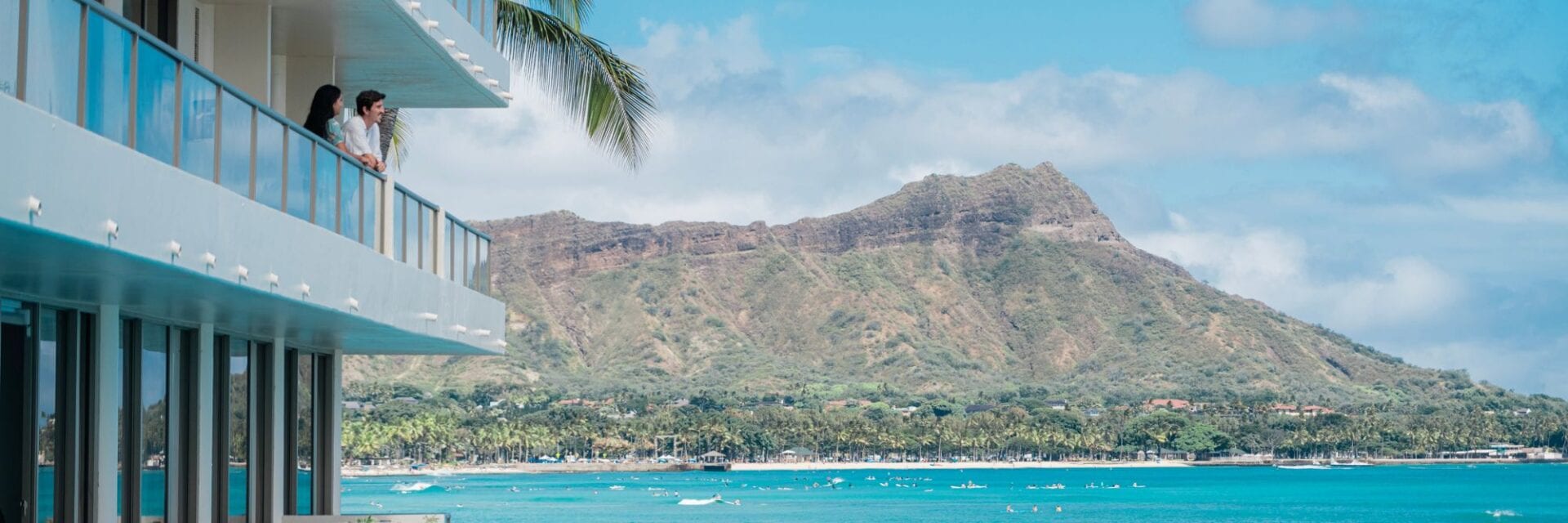 Zwei Menschen stehen auf einem Balkon eines Hotels am Meer und blicken über das türkisfarbene Wasser auf den Diamond Head auf Oʻahu.