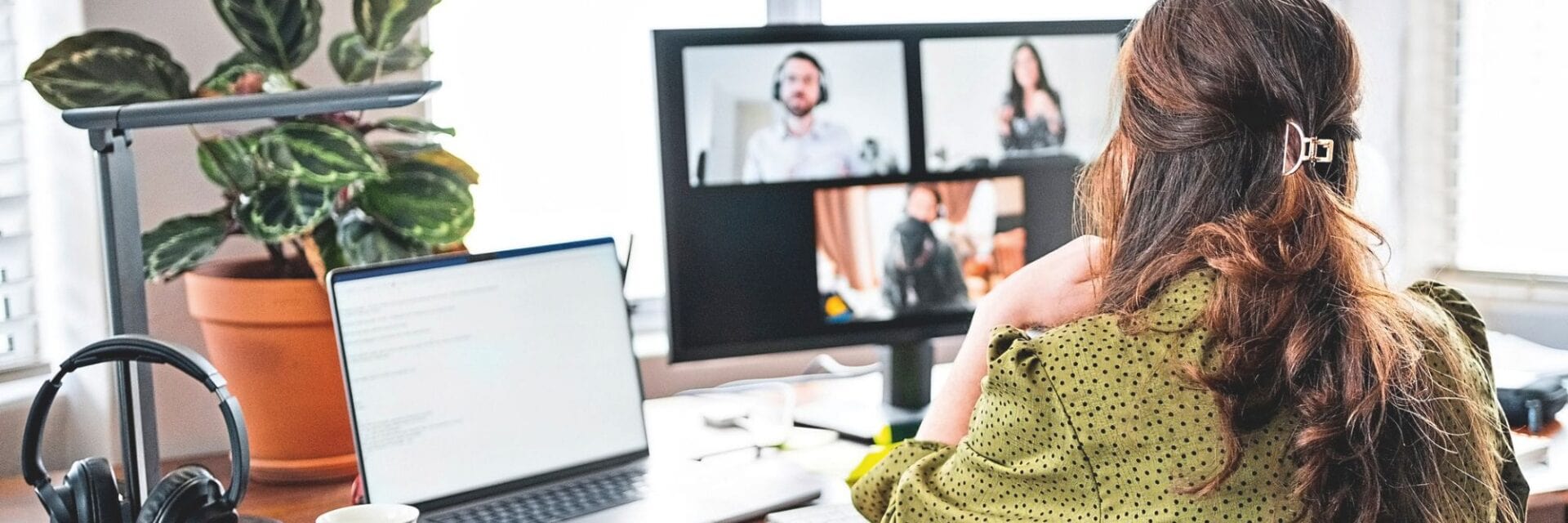 Eine Frau sitzt an einem Schreibtisch im Homeoffice und nimmt an einer Videokonferenz mit mehreren Personen auf dem Bildschirm teil; neben ihr stehen Laptop, Kopfhörer, Kaffeetasse und eine Zimmerpflanze.