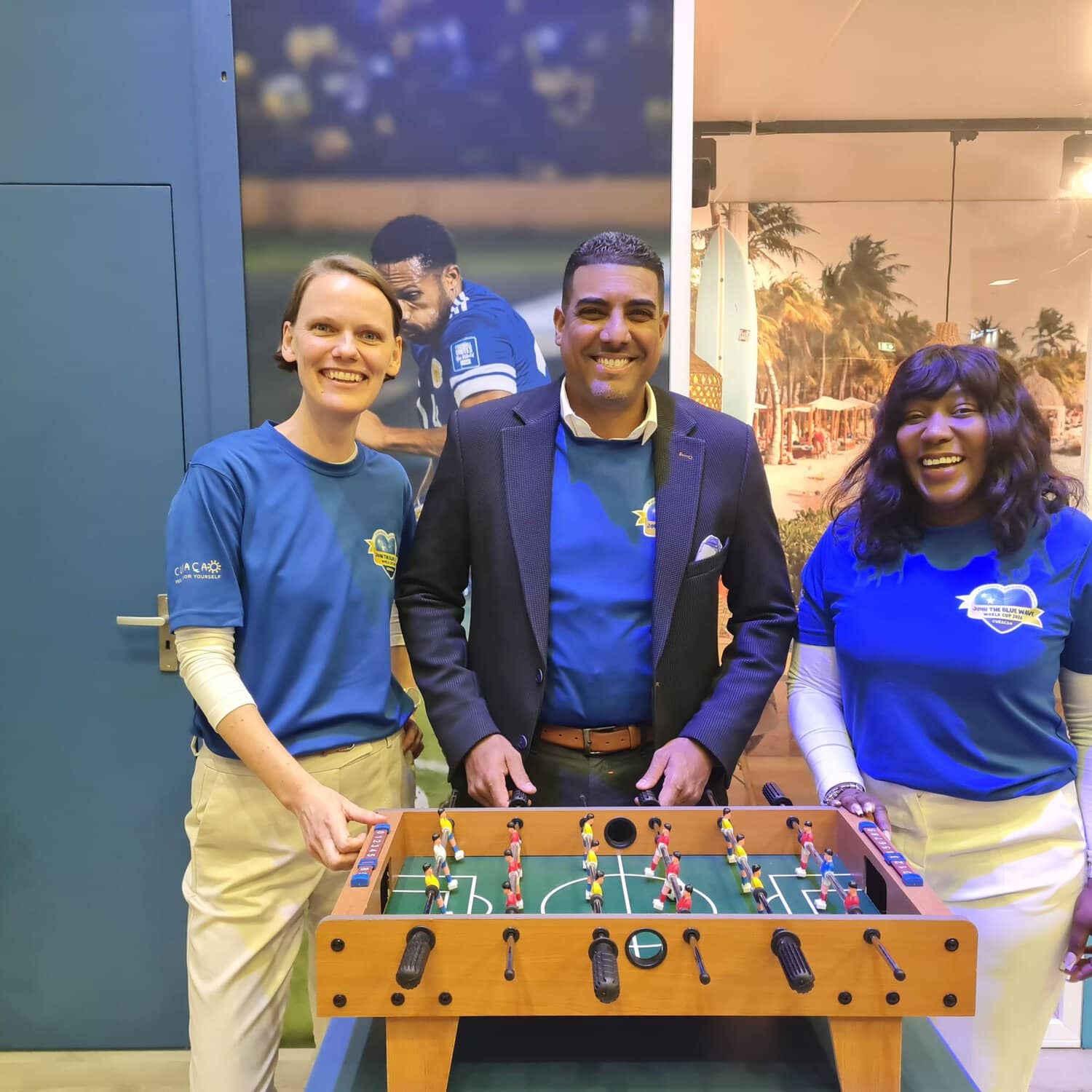 Das Foto zeigt drei Menschen, die an einem Kickertisch stehen, alle in blauem Curacao-Shirt, mit einer tropischen Beach- und Fußballkulisse im Hintergrund.