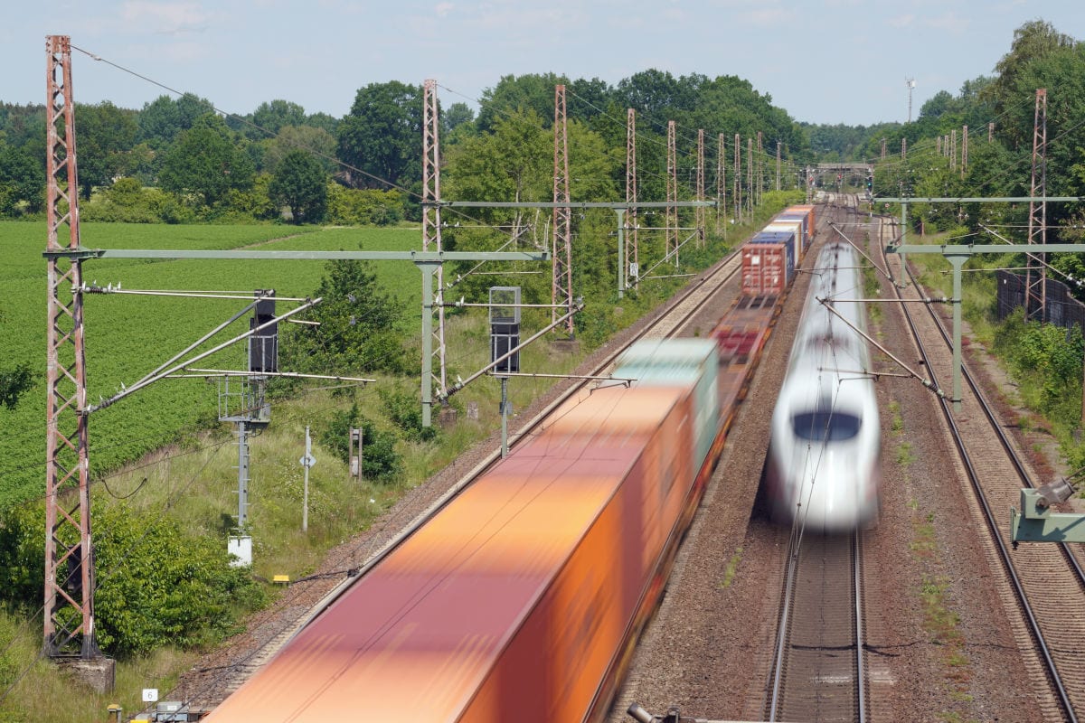 Auf dem Foto sind zwei Züge auf einer Bahnstrecke zwischen Hamburg und Hannover zu sehen: Ein weißer ICE-Hochgeschwindigkeitszug fährt in Richtung Kamera, während ein langer Güterzug mit bunten Containern in die entgegengesetzte Richtung unterwegs ist. Die Gleise verlaufen durch eine grüne Landschaft mit Bäumen und Feldern.