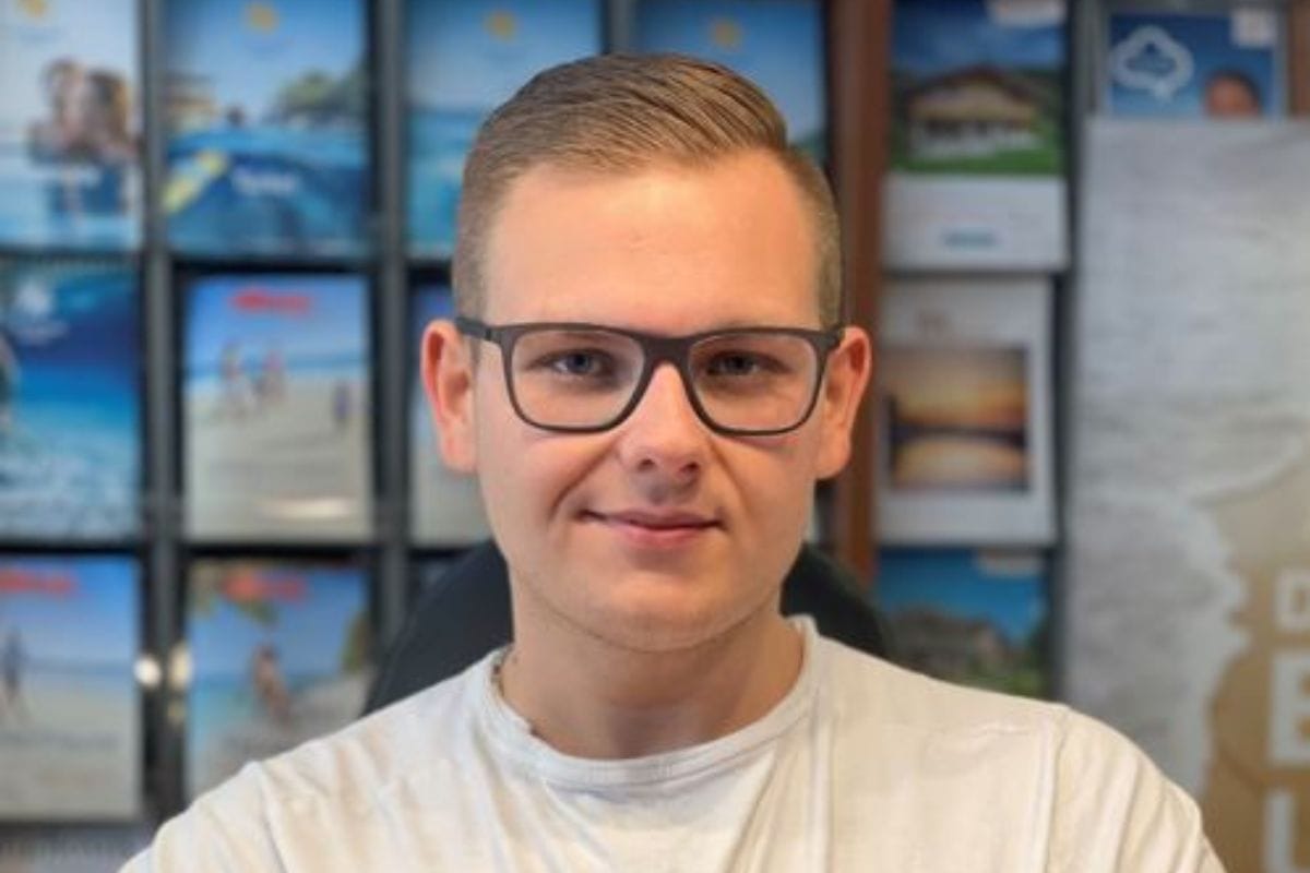 Porträt eines jungen Mannes mit Brille und T-Shirt in einem Reisebüro, vor einer Wand mit Urlaubsprospekten.