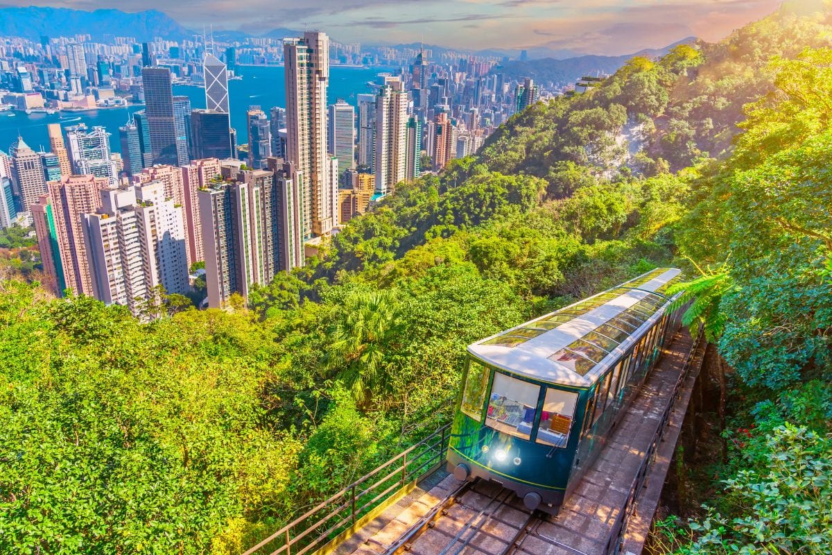 Standseilbahn fährt durch üppig grünen Hang mit Blick auf die Skyline von Hongkong, Hochhäuser und Hafen im Hintergrund bei sonnigem Wetter.