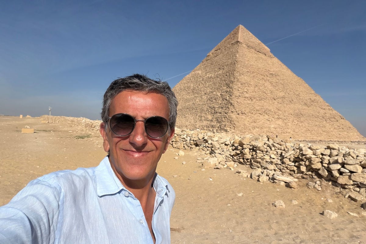 Ein Mann mit kurzen grauen Haaren und Sonnenbrille macht ein Selfie in der Wüste vor einer großen, sandfarbenen Pyramide. Er trägt ein hellblaues Hemd und lächelt in die Kamera. Im Hintergrund sind Steine, flaches Wüstenland und ein klarer blauer Himmel zu sehen.