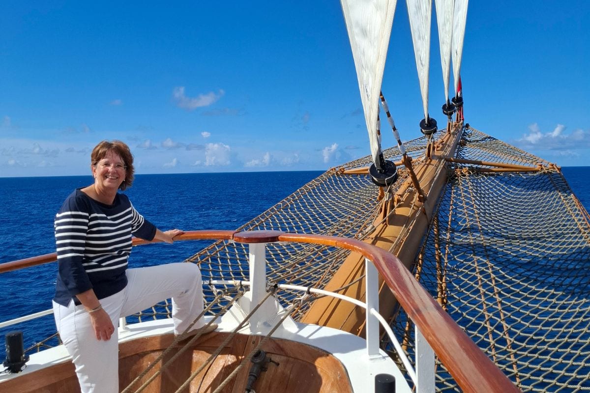 Eine lächelnde Frau steht an Deck eines Segelschiffs, hält sich am Geländer fest und blickt in die Kamera. Im Hintergrund erstrecken sich das offene Meer und ein strahlend blauer Himmel, während die Segel und Netze des Schiffs sichtbar sind.