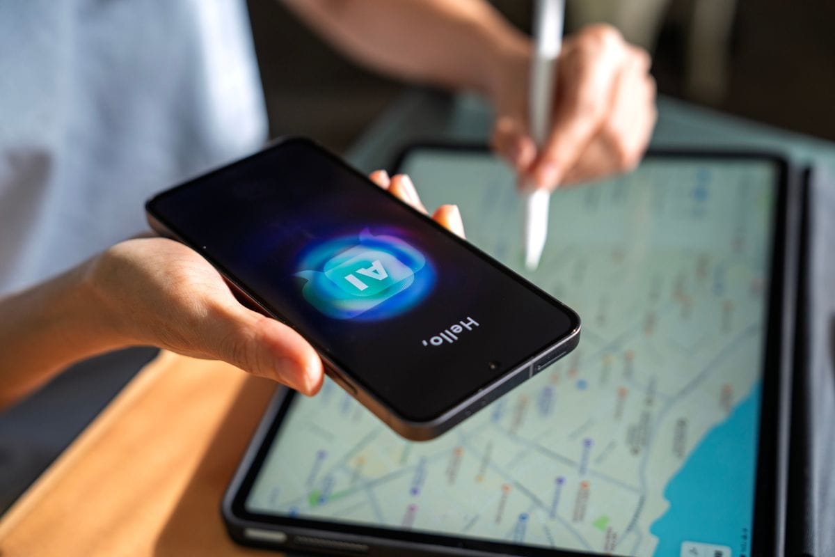 Person hält ein Smartphone mit KI-Icon auf dem Display, während im Hintergrund auf einem Tablet eine digitale Karte mit Routen zu sehen ist.