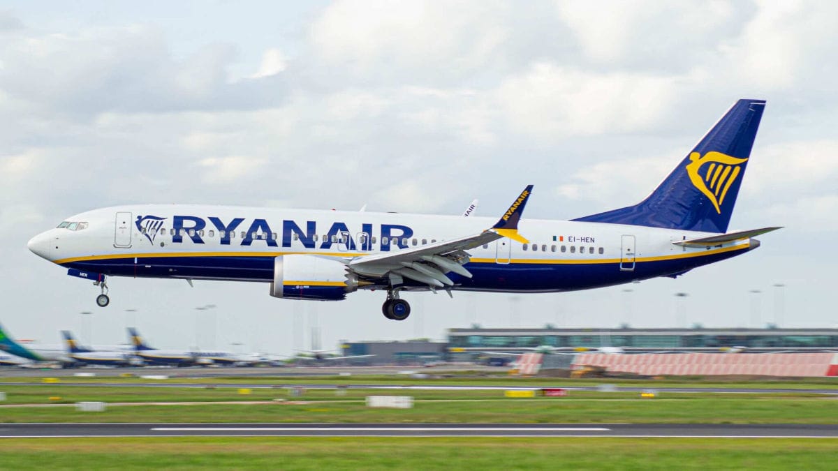 Ein Ryanair-Flugzeug im Landeanflug auf einen Flughafen, mit deutlich sichtbarem Ryanair-Logo und Schriftzug; im Hintergrund sind das Flughafengelände und weitere Flugzeuge zu erkennen.