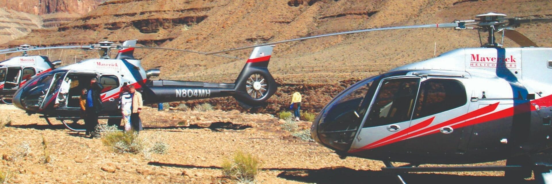 Drei weiße und rote Mark Air Hubschrauber mit „Maverick“-Beschriftung stehen auf dem sandigen Boden am Rand des Grand Canyon. Im Hintergrund ragen die dramatischen, rot-braunen Felsen des Canyons unter klarem Himmel auf. Personen in weißen Shirts sind in der Nähe der Hubschrauber zu sehen.