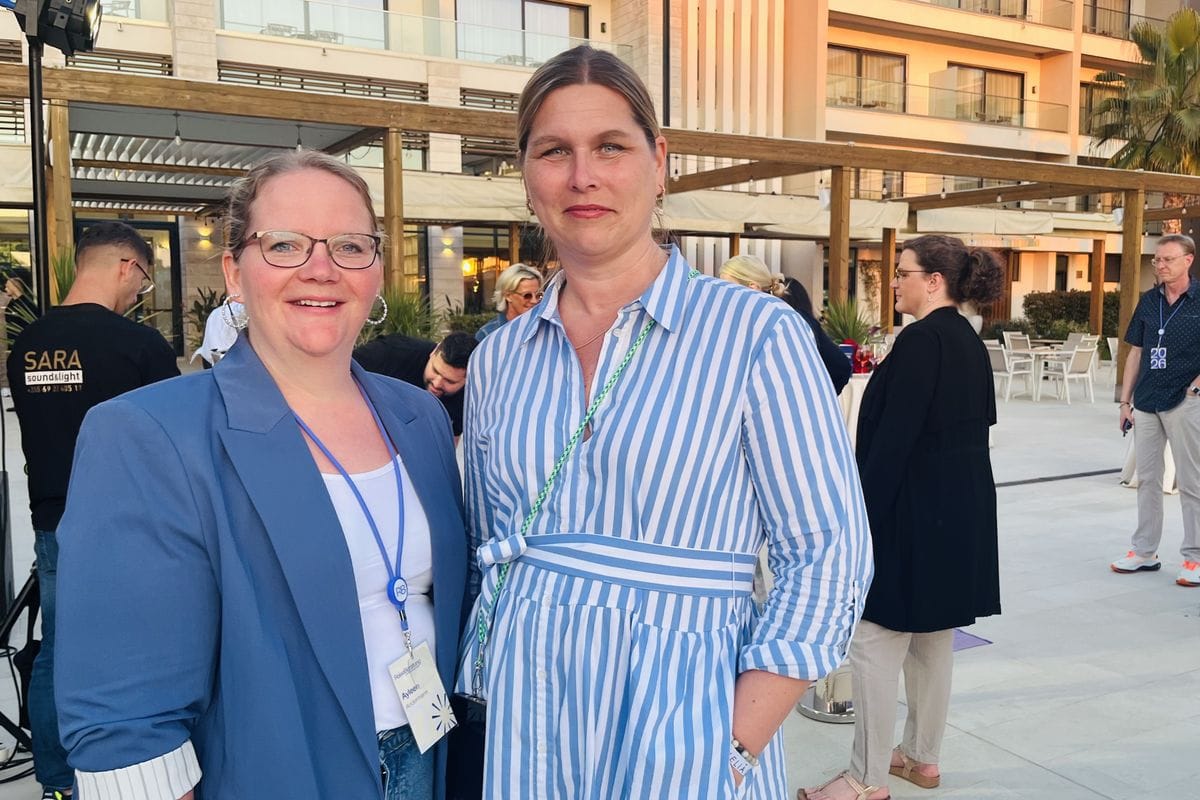 Zwei Frauen stehen vor einem Gebäude mit mehreren Menschen und gucken in die Kamera