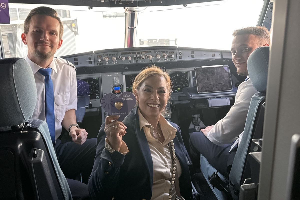 Eine Frau sitzt zwischen zwei Piloten im Cockpit eines Flugzeuges und hält ein Papierherz mit Süße Grüße in die Kamera