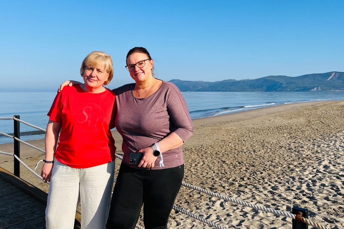 Zwei Frauen stehen auf einem Steg am Strand. Im Hintergrund sind grüne Hügel und das Meer zu sehen
