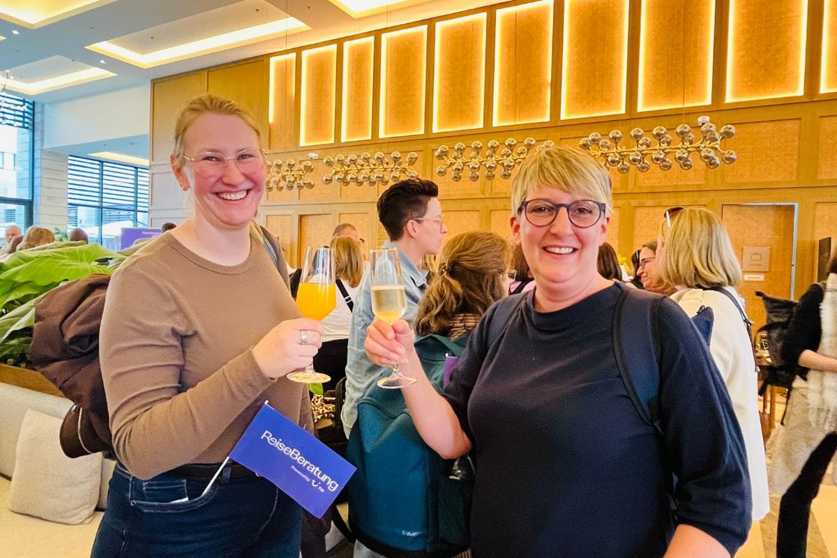 Zwei Frauen stehen in einem gut beleuchteten Innenraum bei einer Veranstaltung und stoßen mit Sektgläsern an. Beide lächeln in die Kamera. Die Frau links trägt eine Brille, ein langärmliges Oberteil und hält zusätzlich eine blaue Mappe mit der Aufschrift „ReiseBeratung“. Die Frau rechts trägt ebenfalls eine Brille und ein dunkles Oberteil. Im Hintergrund sind weitere Gäste zu sehen sowie eine dekorative Wand mit warmem Licht und runden Metallelementen.