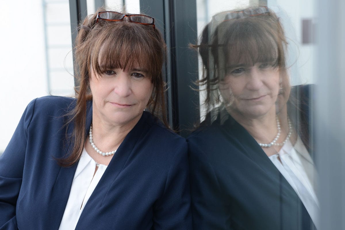 Eine mittelalte Frau mit braunen Haaren und Pony lehnt in einem dunklen Blazer und einer weißen Bluse an einer Glaswand. Eine Lesebrille sitzt auf ihrem Kopf, und sie trägt eine Perlenkette. Ihr nachdenklicher Blick ist leicht nach unten gerichtet, während sich ihr Gesicht deutlich in der Glasscheibe spiegelt.