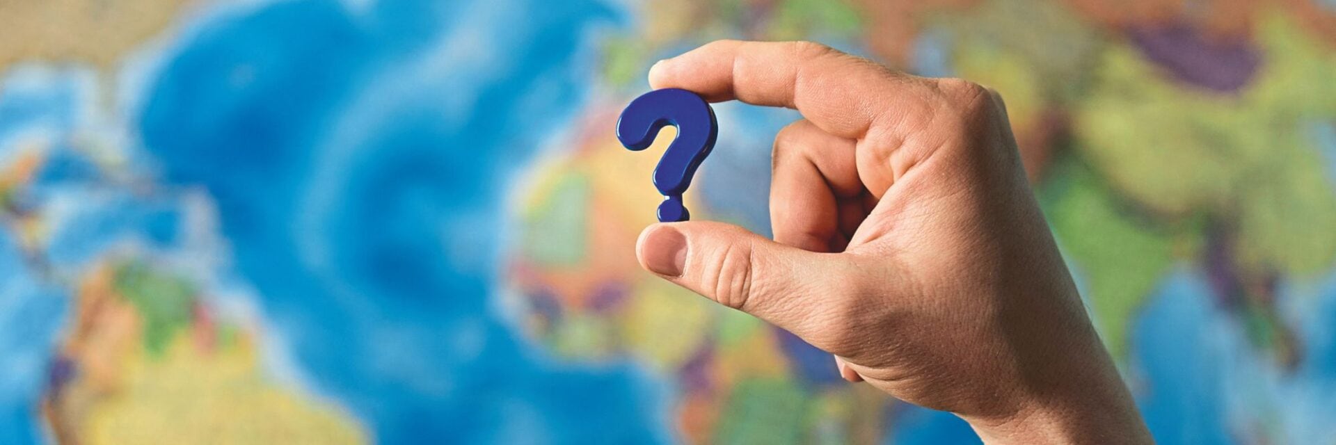 Hand hält ein kleines Fragezeichen vor einer unscharfen Weltkarte im Hintergrund.