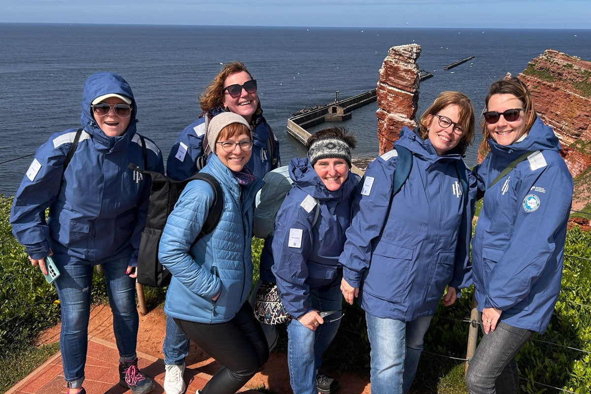 Sechs Personen in blauen Outdoorjacken posieren bei sonnigem Wetter vor der berühmten Felsformation „Lange Anna“ auf Helgoland, mit weitem Blick auf das Meer im Hintergrund. Die Gruppe steht auf einem Weg, umgeben von grüner Vegetation, und genießt den Ausflug auf Deutschlands einziger Hochseeinsel.