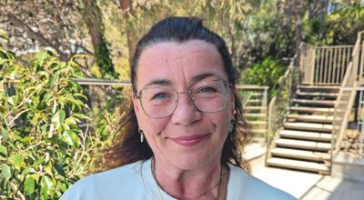 Eine Frau mit zurückgebundenen dunklen Haaren und dunkler Brille steht in einem Garten und schmunzelt in die Kamera.