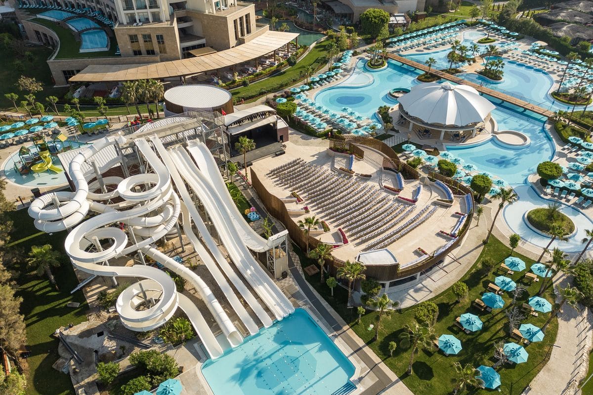 Luftaufnahme eines Freizeitbereichs mit Wasserrutschen, Pools und Liegen in einem Resort, umgeben von Palmen und grünen Rasenflächen.