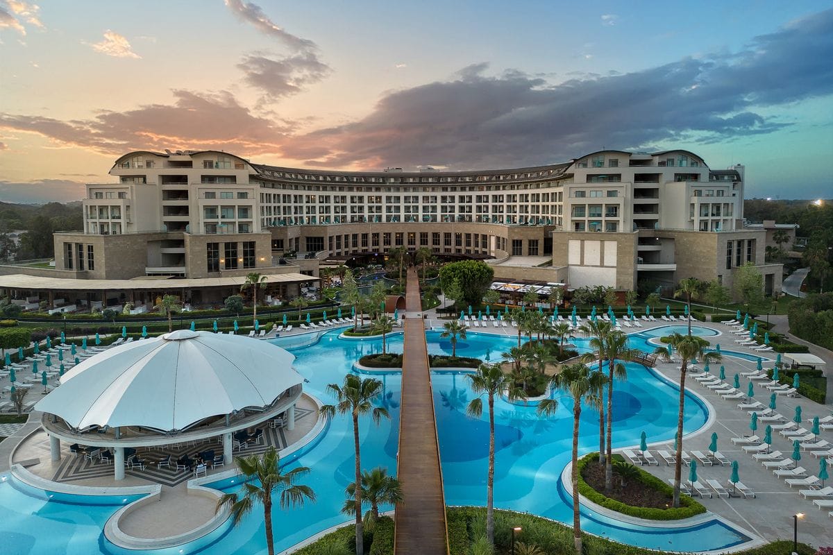 Luxuriöses Hotel mit modernem Design, umgeben von einem Pool und Palmen, unter einem farbenfrohen Himmel bei Sonnenuntergang.