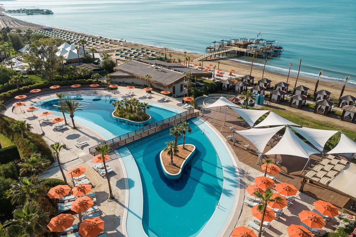Luftaufnahme eines modernen Pools mit Palmen und Liegen, umgeben von orangefarbenen Sonnenschirmen und einer Küstenlandschaft.