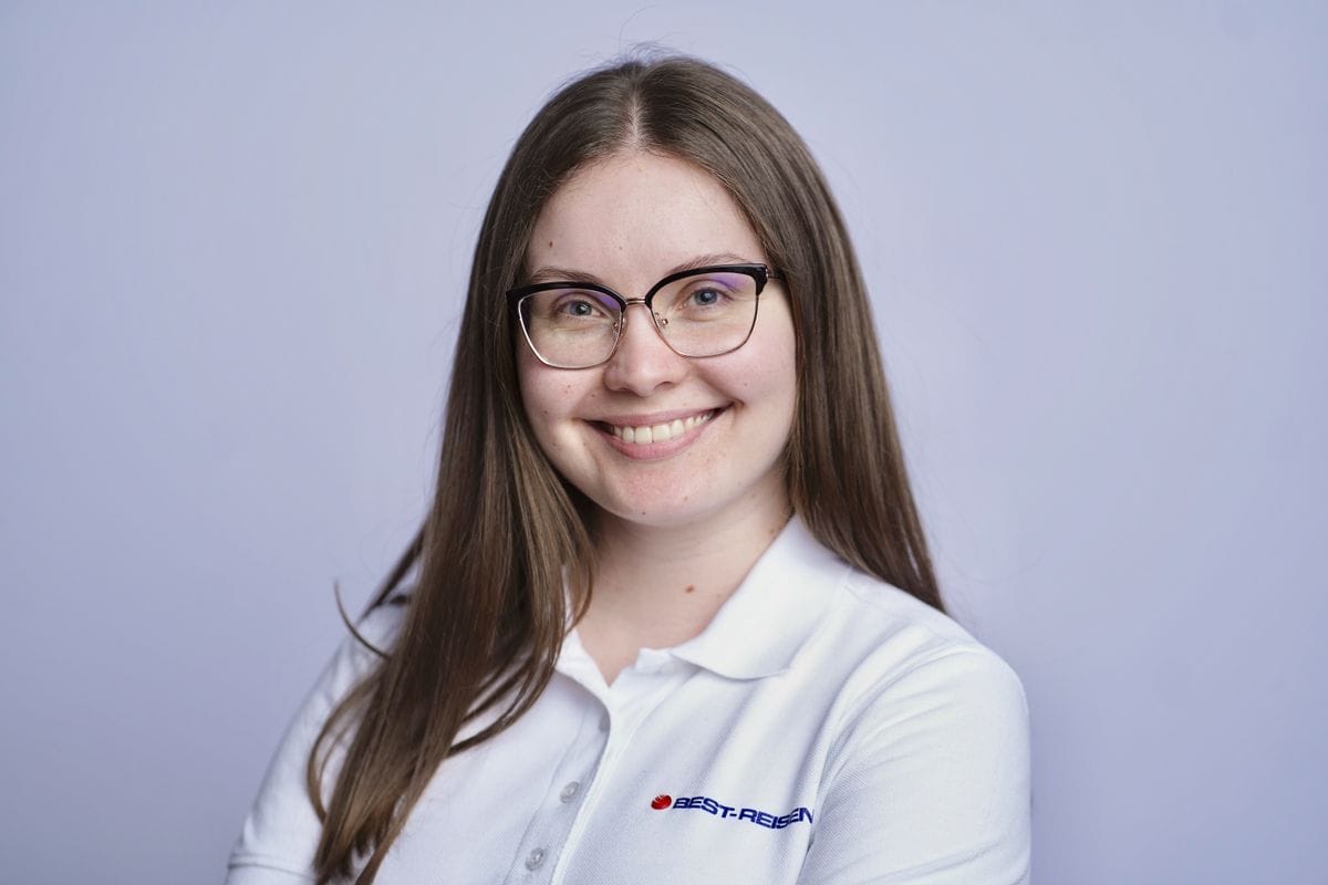 Junge Frau mit langen, braunen Haaren und Brille, die in einem weißen Poloshirt lächelt, vor einem einfarbigen Hintergrund.