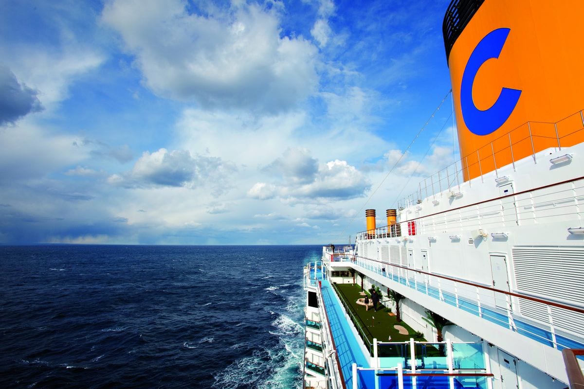 Seitenansicht eines Costa-Kreuzfahrtschiffs auf offenem Meer unter dramatischem, wolkigem Himmel, mit orangefarbenem Schornstein und blauem Logo.
