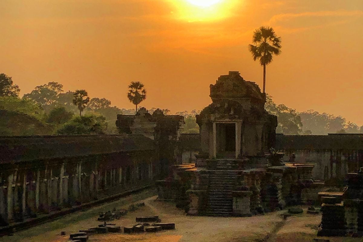Die Ruinen eines Tempels liegen im warmen Licht des Sonnenuntergangs, während die tief stehende Sonne den Himmel orange färbt und Palmen silhouettiert.