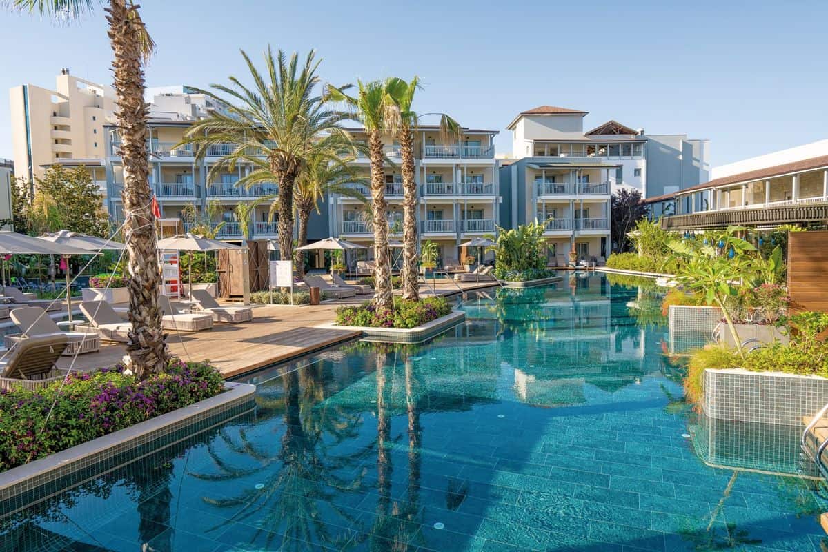 Ein luxuriöser Hotelpool umgeben von Palmen und Liegen, mit modernen Gebäuden im Hintergrund und klarem blauen Himmel.